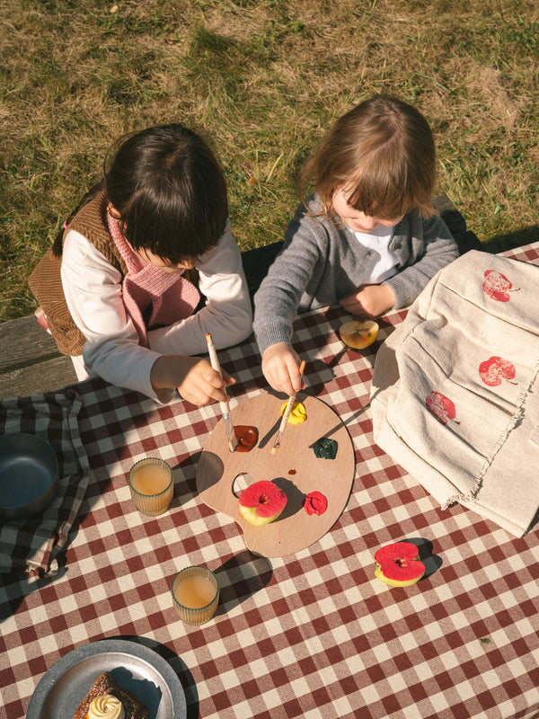 Evento Apple Stamping, dos niños estampan mitades de manzana sobre un cesto textil en una mesa de picnic de cuadros