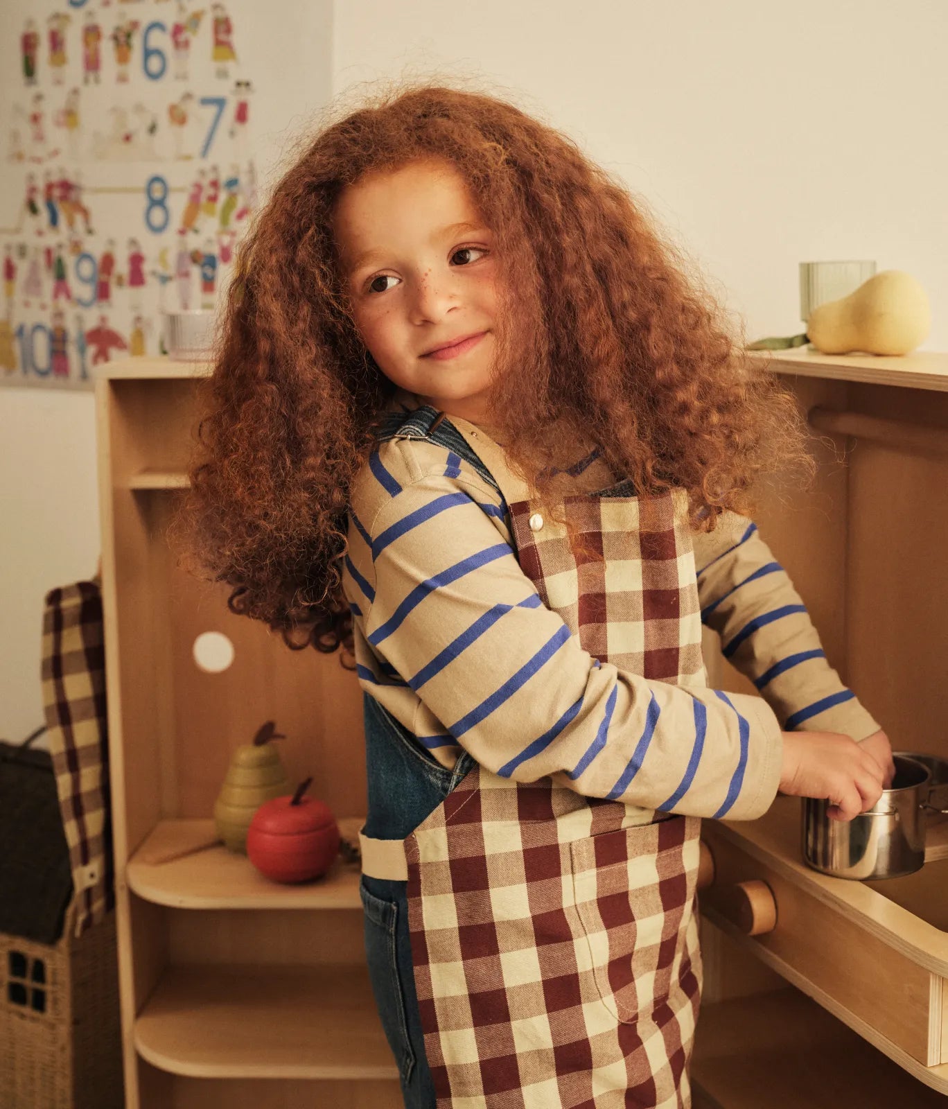 Collection Kids Autumn Winter 2026, enfant en tablier à carreaux tenant une casserole devant une cuisine de jeu